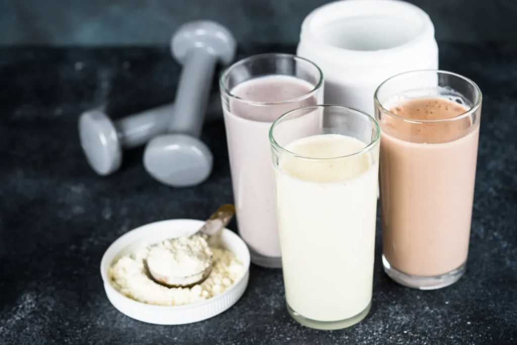 Verschiedene Proteinshakes in Gläsern mit Hanteln und Proteinpulver als Fitnessnahrung.