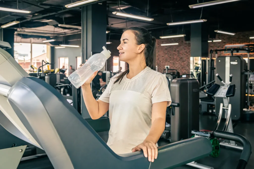 Junge Frau trinkt Wasser auf einem Laufband im Fitnessstudio während ihres HIIT-Trainings.