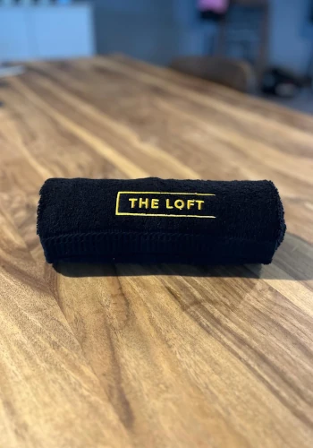 The Loft Handtuch – Schwarzes Handtuch mit gelbem "The Loft"-Logo, zusammengerollt auf einem Holztisch.