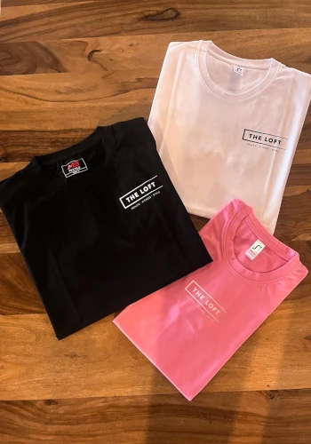 The Loft T-Shirts – Drei T-Shirts in Schwarz, Weiß und Pink mit "The Loft"-Branding, auf einem Holztisch liegend.