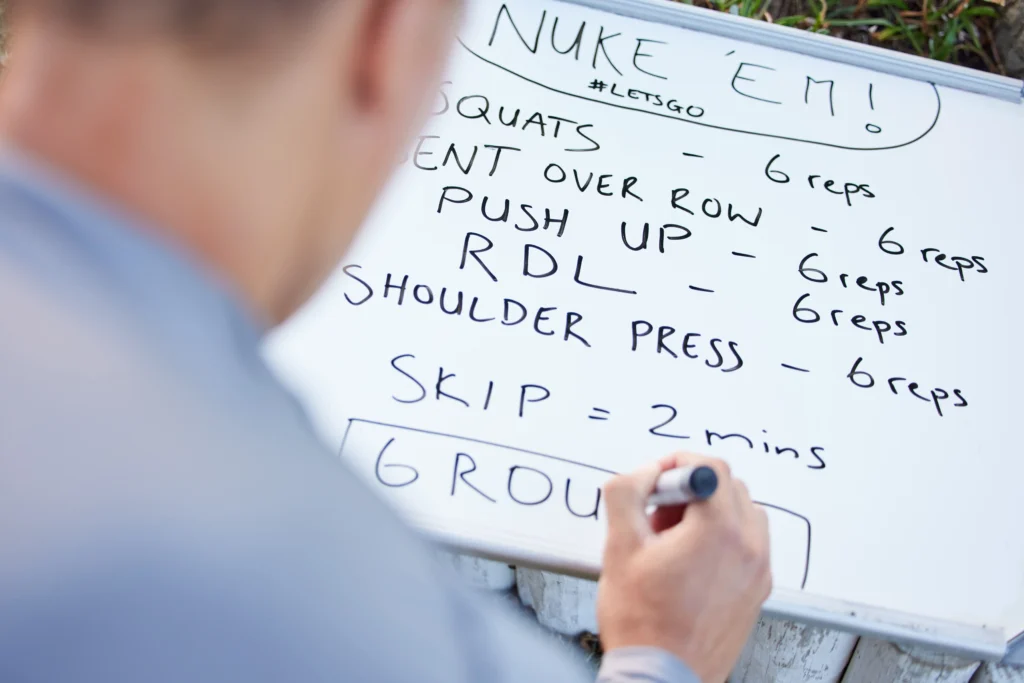 Trainer schreibt einen Trainingsplan auf ein Whiteboard mit Übungen wie Squats, Push-ups und Shoulder Press – ideal für Muskelaufbau und Fettverbrennung
