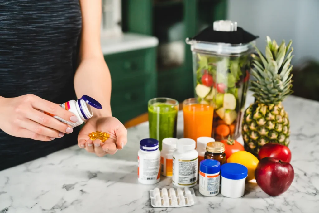 Frau mit Nahrungsergänzungsmitteln in der Hand vor frischen Früchten, Säften und einem Mixer – Symbolbild für gesunde Ernährung und Supplement-Einsatz im Sommertraining