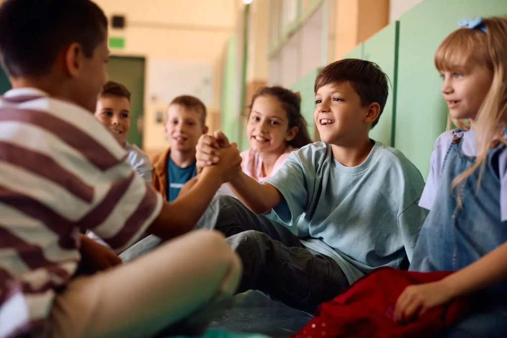 Eine Gruppe von Kindern sitzt im Kreis, zwei Jungs drücken sich spielerisch die Hände – Gemeinschaft, Vertrauen und Selbstvertrauen im Fokus.