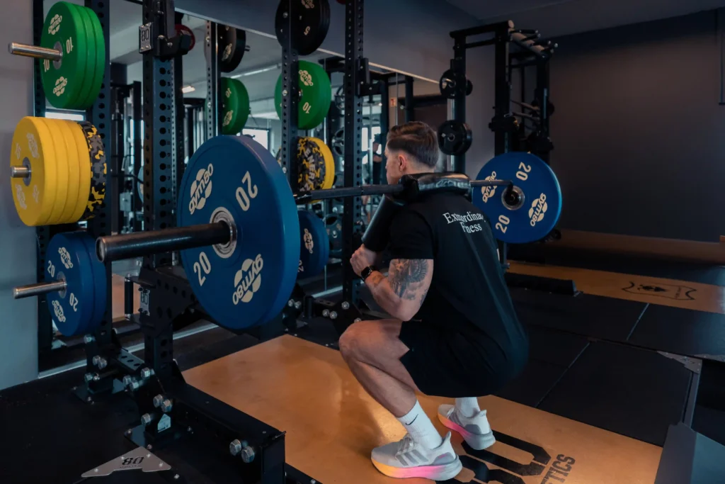 Muskulöser Mann mit tätowiertem Arm führt eine Kniebeuge mit einer beladenen Langhantel an einer Squat Rack-Station in einem modernen Fitnessstudio aus.