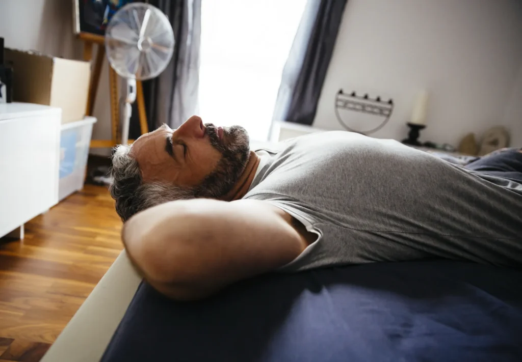 Mann liegt entspannt mit geschlossenen Augen im Bett, um sich nach dem Training durch Schlaf zu regenerieren.