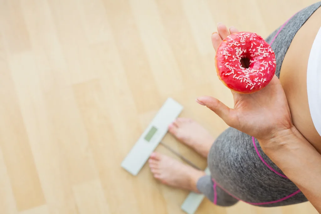 Frau mit Donut neben einer Waage als Symbol für Heißhunger, Ernährung und Abnehmen
