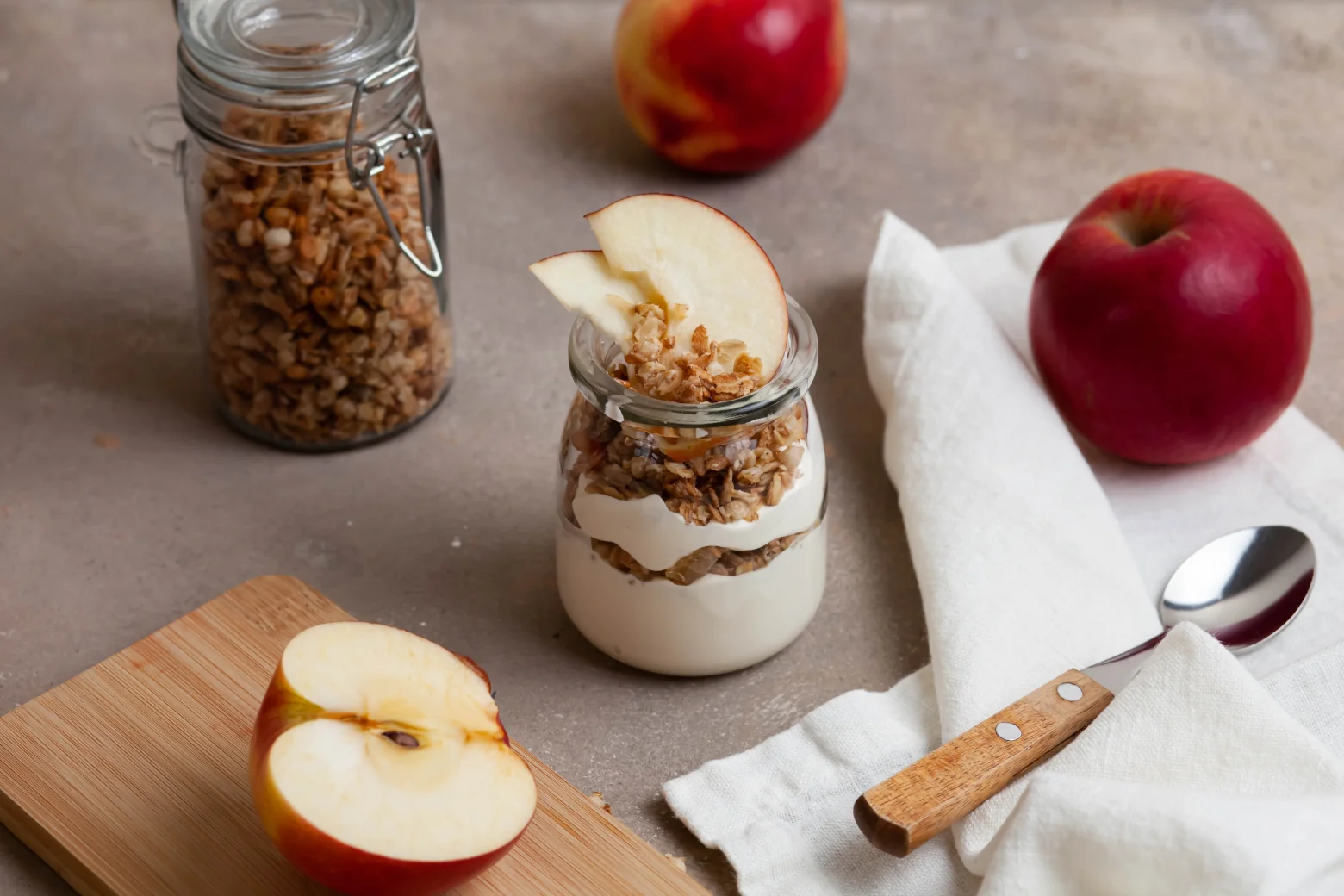 Joghurt und Granola in einem Glas geschichtet und mit Apfelscheiben garniert, daneben frische Äpfel.
